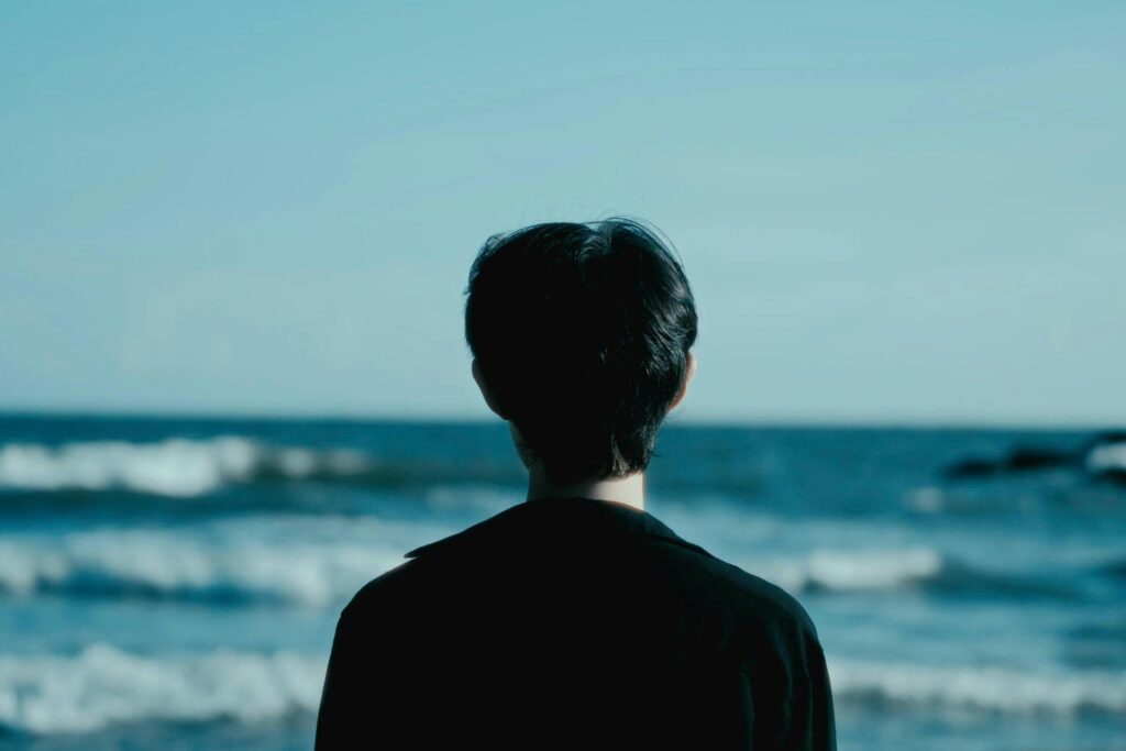 Person standing by the ocean, looking out at the waves under a clear blue sky.